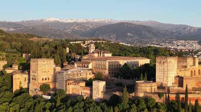 Drone aerial view of the Alhambra Palace in Granada, Spain glowing in golden hour light with the snow-capped Sierra Nevada mountains stretching across the horizon