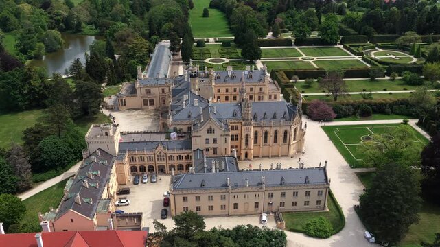 High-altitude aerial view of Lednice Castle with its full grounds, formal hedged gardens, and surrounding parkland with a lake visible, Moravia, Czech Republic.