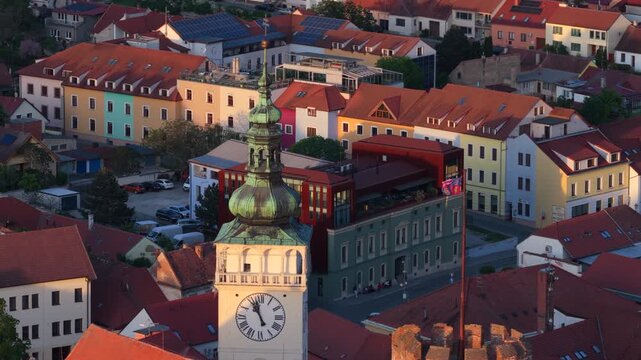 Aerial drone view of a baroque clock tower with green copper onion dome rising above a small-town square surrounded by colourful buildings in warm sunset light, Mikulov, Moravia, Czech Republic.
