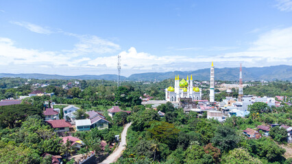 Fototapeta premium Aerial drone view of the Great Mosque of Parepare cityscape with green yellow domes, residential houses, and lush green trees under clear blue sky in South Sulawesi, Indonesia.