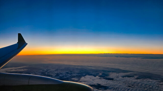 Airplane wing and engine seen from the window, soaring above a fluffy cloud layer, with a colorful gradient horizon and deep blue sky reflecting the beautiful light of dusk or dawn