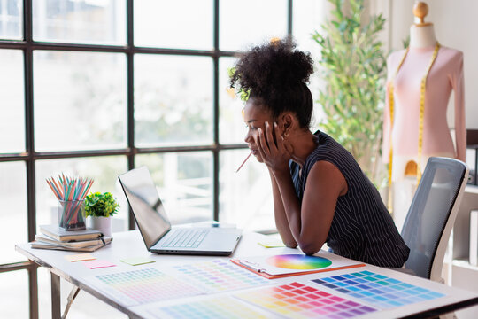 African american fashion designer sitting at a desk with a laptop and color swatches, contemplating designs and experiencing feelings of stress and contemplation while working in her studio