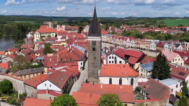 Aerial drone view of a tall Gothic clock tower rising above a historic Czech town square with colorful arcaded townhouses, church towers, and rolling countryside beyond, Telc, Czech Republic