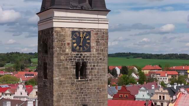 Aerial drone view of a medieval stone clock tower with a Roman numeral clock and a church steeple rising above red-roofed buildings with green hills in the background, Telc, Czech Republic