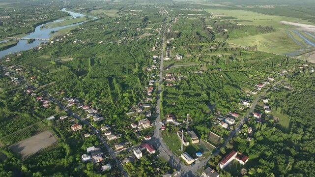 High angle drone shot of Anaklia village surrounded by lush green forests, agricultural fields, and a winding river under bright daylight.