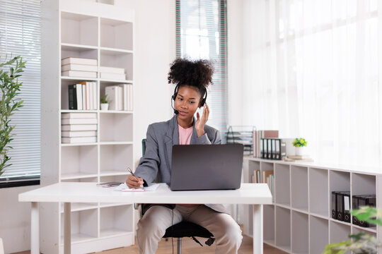 African american businesswoman wearing headset, providing online customer service while working remote from home office, consulting laptop and writing notes