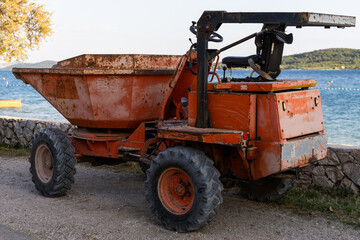 An old rusty orange site dumper truck parked on a gravel path near a stone wall by the sea, construction machinery in a coastal area. © Vinona_Forever