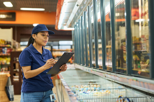 Hispanic young woman working in supermarket, checking frozen food inventory on clipboard near freezer aisle. Useful for retail staffing, grocery operations, supply chain marketing
