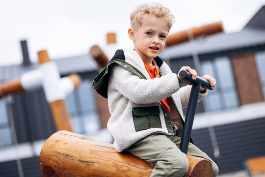 Blond child enjoying outdoor play, holding handlebars of a wooden rocking toy with a happy expression, enjoying childhood