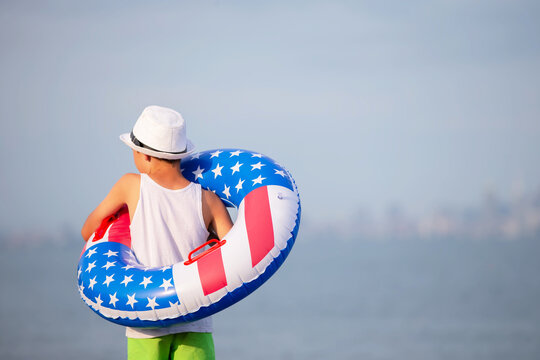 A child in a white hat stands on the shore, holding a round float