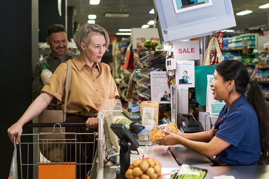 Hispanic young woman working at supermarket checkout and adult shopping for groceries, supporting retail service and consumer purchase marketing campaigns
