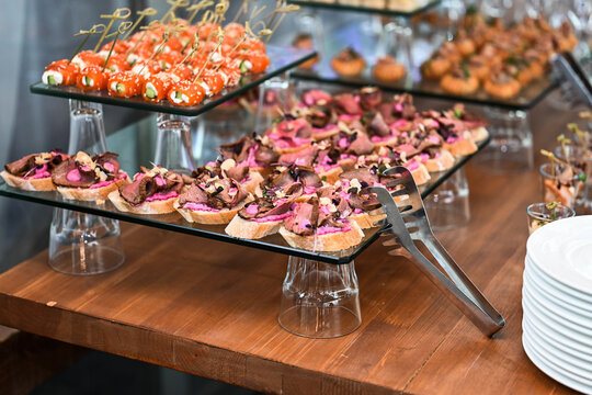 Gourmet buffet table with roast beef bruschetta appetizers on glass stands and metal food serving tongs on wooden counter