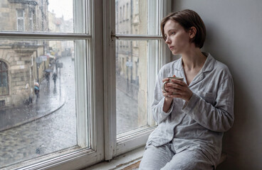 Fototapeta premium A sad girl in pajamas is sitting on the windowsill and drinking tea