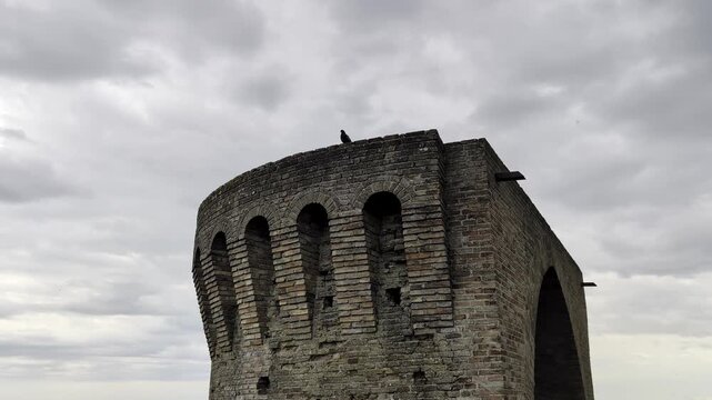 Torre storica in pietra con archi verticali e cielo grigio compatto, per un&rsquo;atmosfera medievale e suggestiva tipica dei borghi fortificati marchigiani