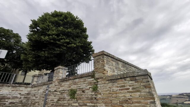 Scorcio di Offagna con muro in pietra, ringhiera panoramica e albero imponente sotto un cielo nuvoloso