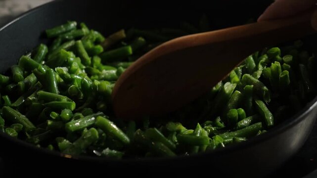 Hand with wooden spoon stirring green bean pods in pan. Preparing plant based vegetarian meal