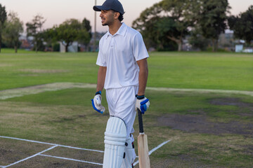 Obraz premium Indian male cricketer standing on pitch at dusk holding bat wearing pads gloves cap