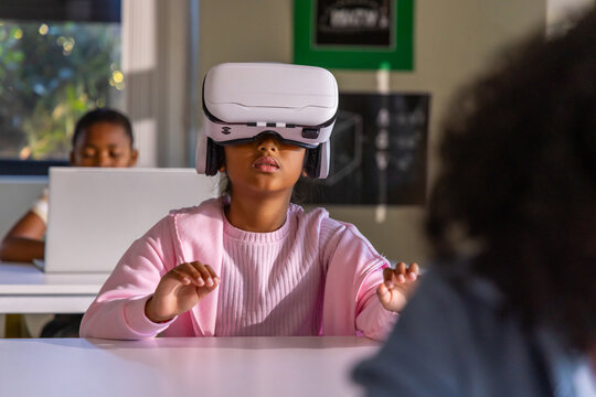 African American classmates at desks watching girl wearing white VR headset using hands with laptop