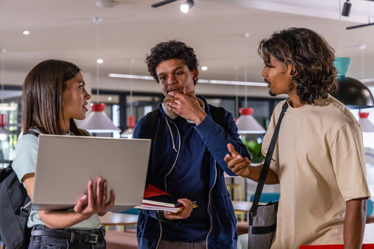 Diverse students standing in study lounge, sharing laptop and notebooks, wearing casual backpacks