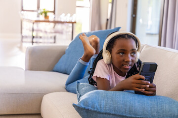 Fototapeta premium School-age African American girl lying on sofa at home wearing white headphones holding device