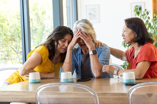 Diverse female friends sitting at wooden table comforting senior woman, three teal mugs, smartphone