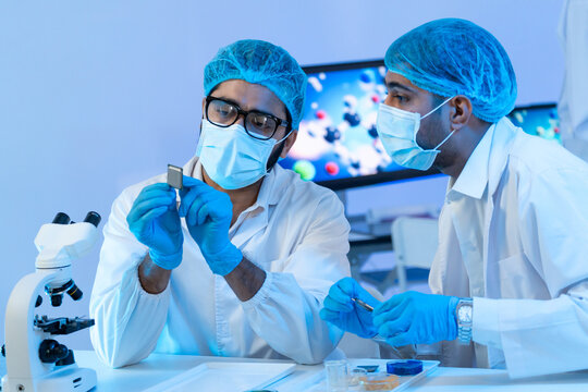 Two male engineers in protective gear carefully examining a microchip semiconductor component together in a high-tech modern laboratory setting.