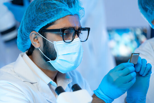 Close-up portrait of a focused male technician wearing safety glasses, face mask, and gloves carefully inspecting a semiconductor microchip in a modern high-tech laboratory.