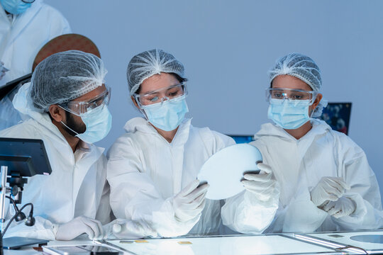 Diverse team of engineers wearing full protective suits and masks carefully inspects a semiconductor silicon wafer over a light table in a high-tech laboratory.