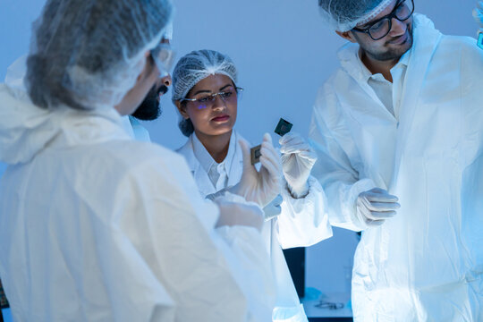 Diverse team of engineers in protective cleanroom suits hold and examine semiconductor microchips together in a modern high-tech laboratory.