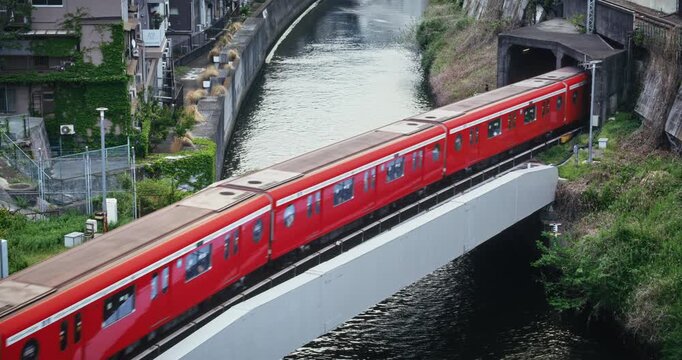 Bright red train exiting tunnel and crossing bridge over river in Japan - high, locked off shot