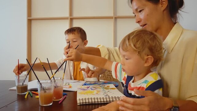 Young creative mother and her two little sons having fun painting with watercolors at home, engaging in a family artistic activity that promotes bonding and creativity for the children