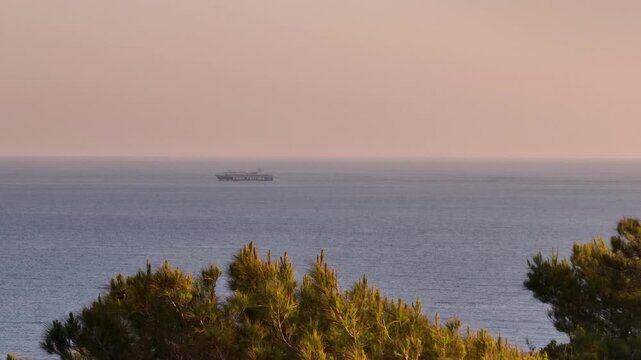Aerial view past coastal pine foliage to a distant cargo ship crossing a calm Mediterranean sea at sunset