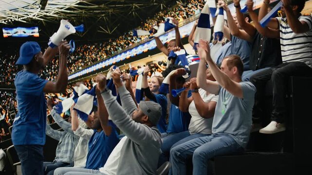 Blue team football capo standing above crowd with megaphone, leading passionate supporter section during energetic stadium chant, organized fans producing powerful support chants with crowd energy.