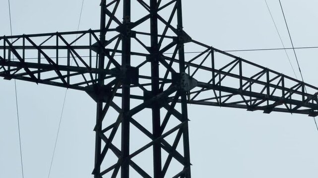 The dark metal beams of a large electricity pylon interlock against a light gray sky. This massive industrial structure creates an impressive feeling of silent strength and stability.