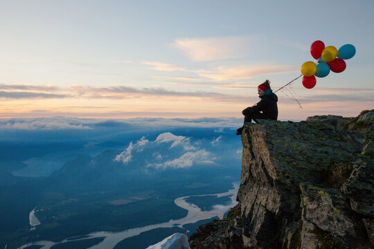 Hiker sits with colorful balloons on a mountain peak