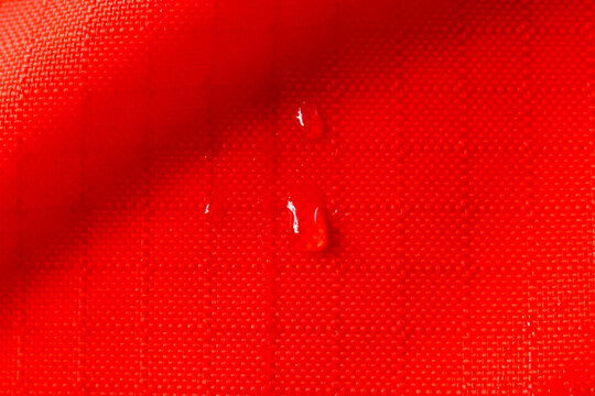 Vibrant red fabric background shows waterproof textile material with clean ripstop texture, featuring fresh water droplet beads reflecting bold energy