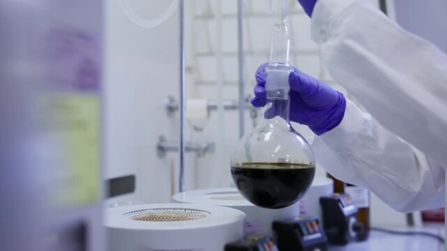Scientist pouring dark oil fuel into round flask on hotplate in factory laboratory.