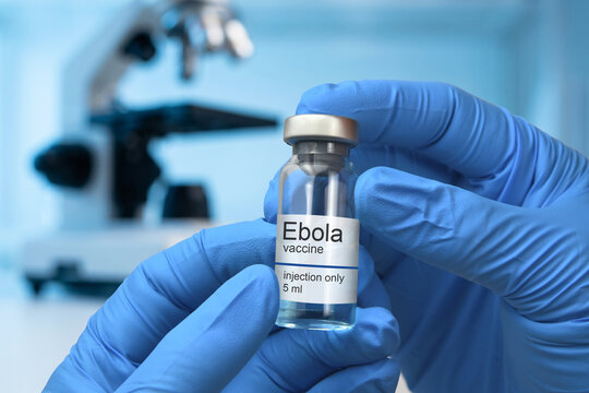 A researcher in blue gloves holds a vial labeled Ebola vaccine in a laboratory setting blurred in the background