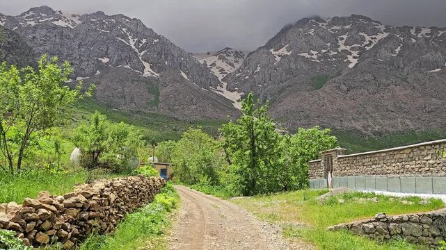 Gravel road leads between a stone wall and lush green spring trees toward snow-streaked rocky mountains under a heavy stormy sky.
📍 Hawraman Takht, Kurdistan, Iran 