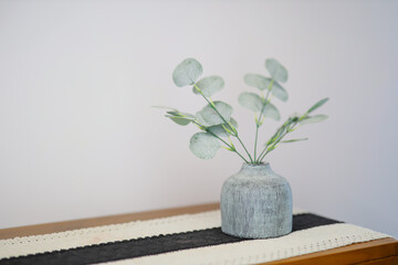 Eucalyptus Branches. A minimalist interior decoration featuring green eucalyptus branches in a textured grey ceramic vase, placed on a wooden table with a fabric runner. © Rijjal