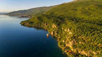 Obraz premium Aerial drone panorama of a scenic lakeside village on Lake Ohrid, North Macedonia. Pristine blue shoreline bordered by lush green mountains and red-roofed houses