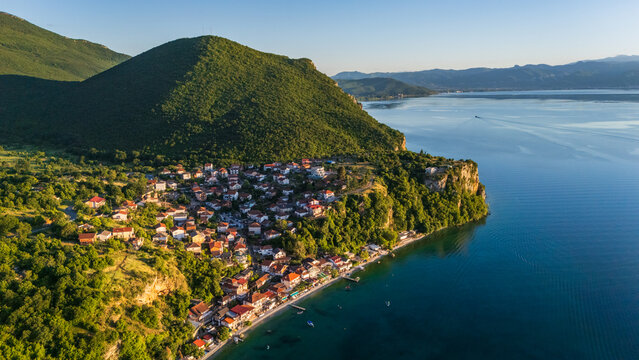 Aerial drone panorama of a scenic lakeside village on Lake Ohrid, North Macedonia. Pristine blue shoreline bordered by lush green mountains and red-roofed houses