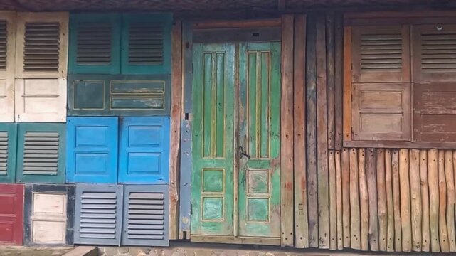 Colorful doors and shutters line a rustic wooden wall. Weathered paint adds character to each panel. Vintage green door stands central. Stone steps lead to the entrance.
