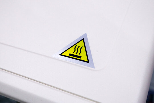 Close up of a yellow triangular hot surface warning sticker on the white metal body of a kitchen appliance