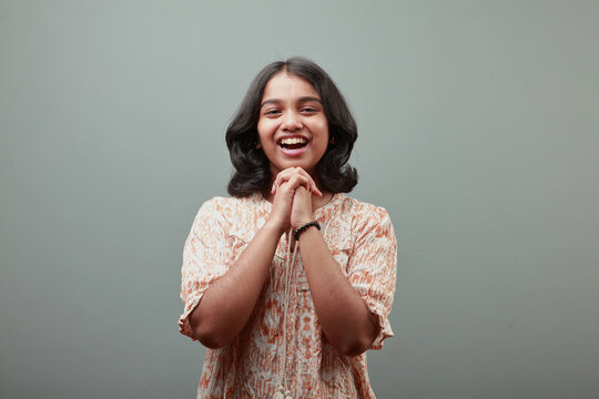Smiling Indian teenage girl with hands clasped under chin posing against a gray studio background, expressing happiness, confidence and positivity
