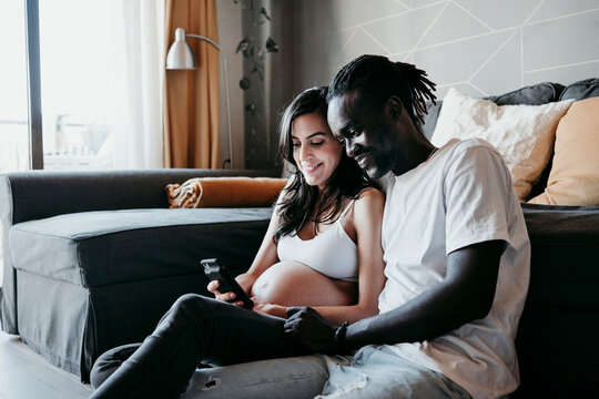 Young man with pregnant woman using smart phone while sitting in living room at home