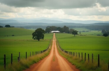 Dirt road leads to farmhouse in rolling green hills and distant mountains under a cloudy sky. Rural Australian landscape offers scenic beauty and tranquility.