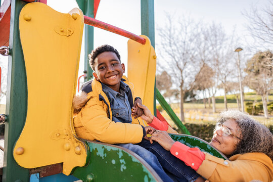 Playful son sitting on slide by grandmother on playground