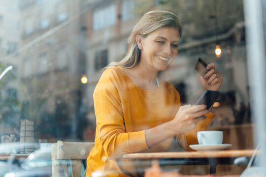 Happy woman using smart phone with credit card in cafe seen through glass