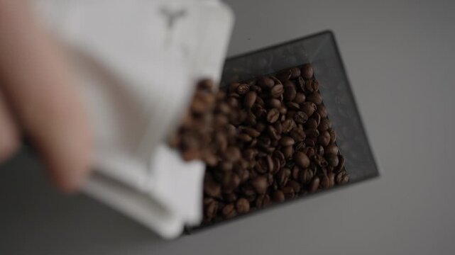 Close-up shot of person hand pouring freshly roasted coffee beans from white paper bag into gray container, preparing them for grinding to make invigorating drink. Shooting in slow motion